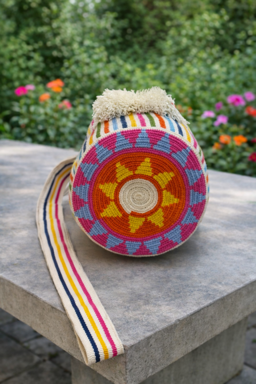 Handgemaakte Wayuu tas Catalina Sofia Medium van LolaGrace met kleurrijk bohemian patroon op een betonnen tafel in een bloeiende tuin.