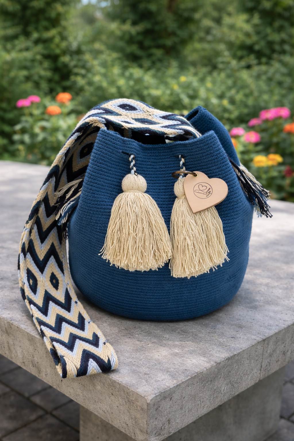 Handgemaakte Wayuu tas Gabriela María Large van LolaGrace in diepblauw met beige kwastjes op een betonnen tafel in een bloeiende tuin.