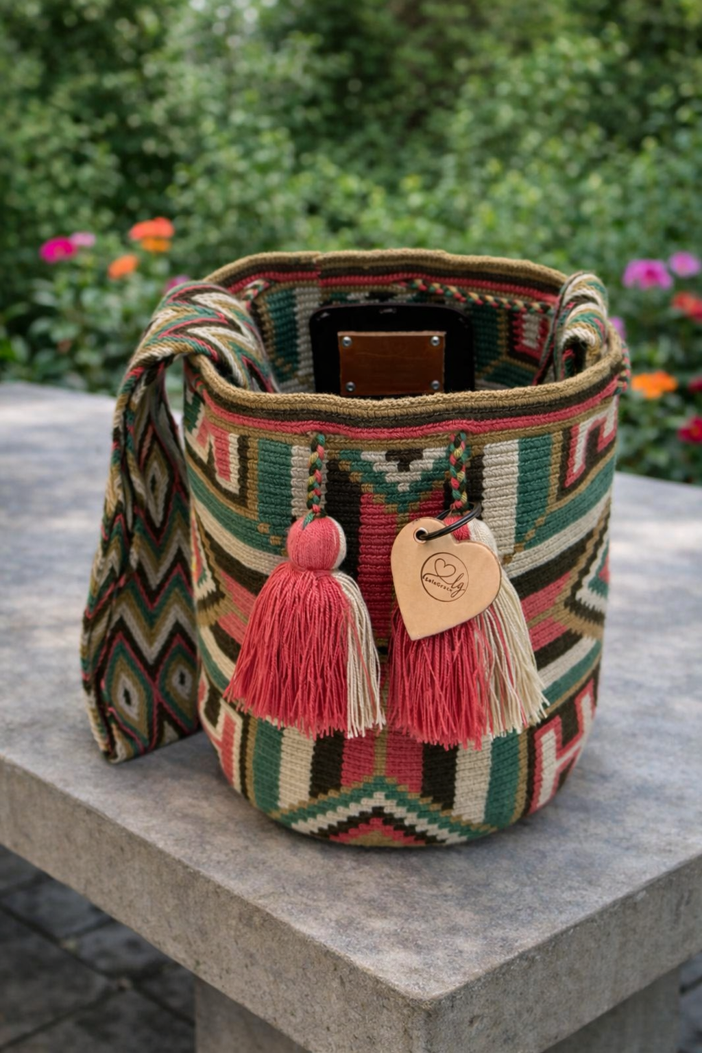 Handgemaakte Wayuu tas Luz Elena Large van LolaGrace met groen en roze traditioneel patroon op een betonnen tafel in een bloeiende tuin.