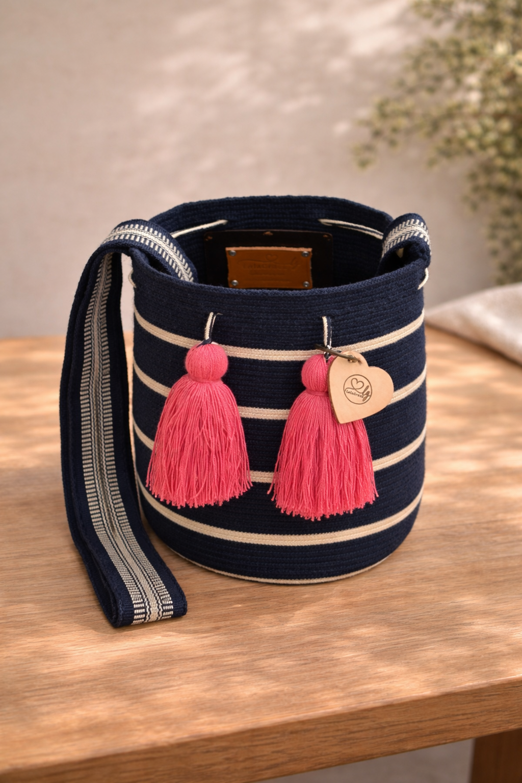 Open Melany Medium Wayuu mochila tas van LolaGrace op houten tafel, handgemaakt door de Wayuu gemeenschap in La Guajira, Colombia, met donkerblauwe strepen en roze kwasten.