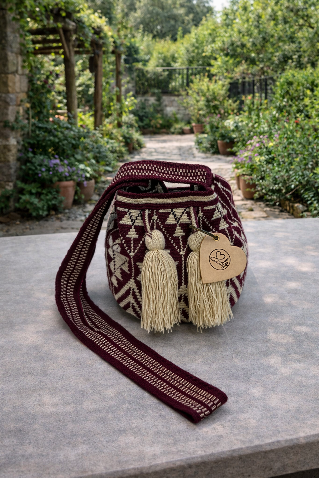 Ambachtelijk geweven Wayuu tas Rosa Maria in diep bordeaux met paarse ondertoon, gepresenteerd in een sfeervolle groene tuinomgeving