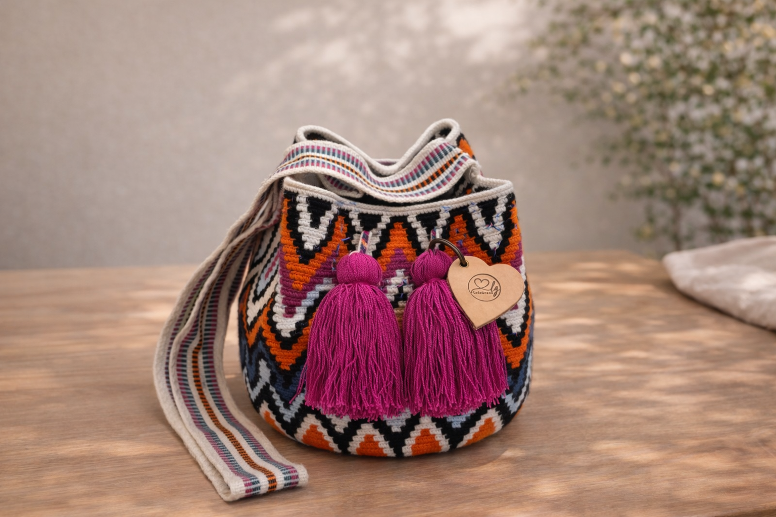 Rosaura Medium Wayuu mochila tas van LolaGrace op houten tafel, handgemaakt door de Wayuu gemeenschap in La Guajira, Colombia, met kleurrijk bohemian patroon en roze kwasten.