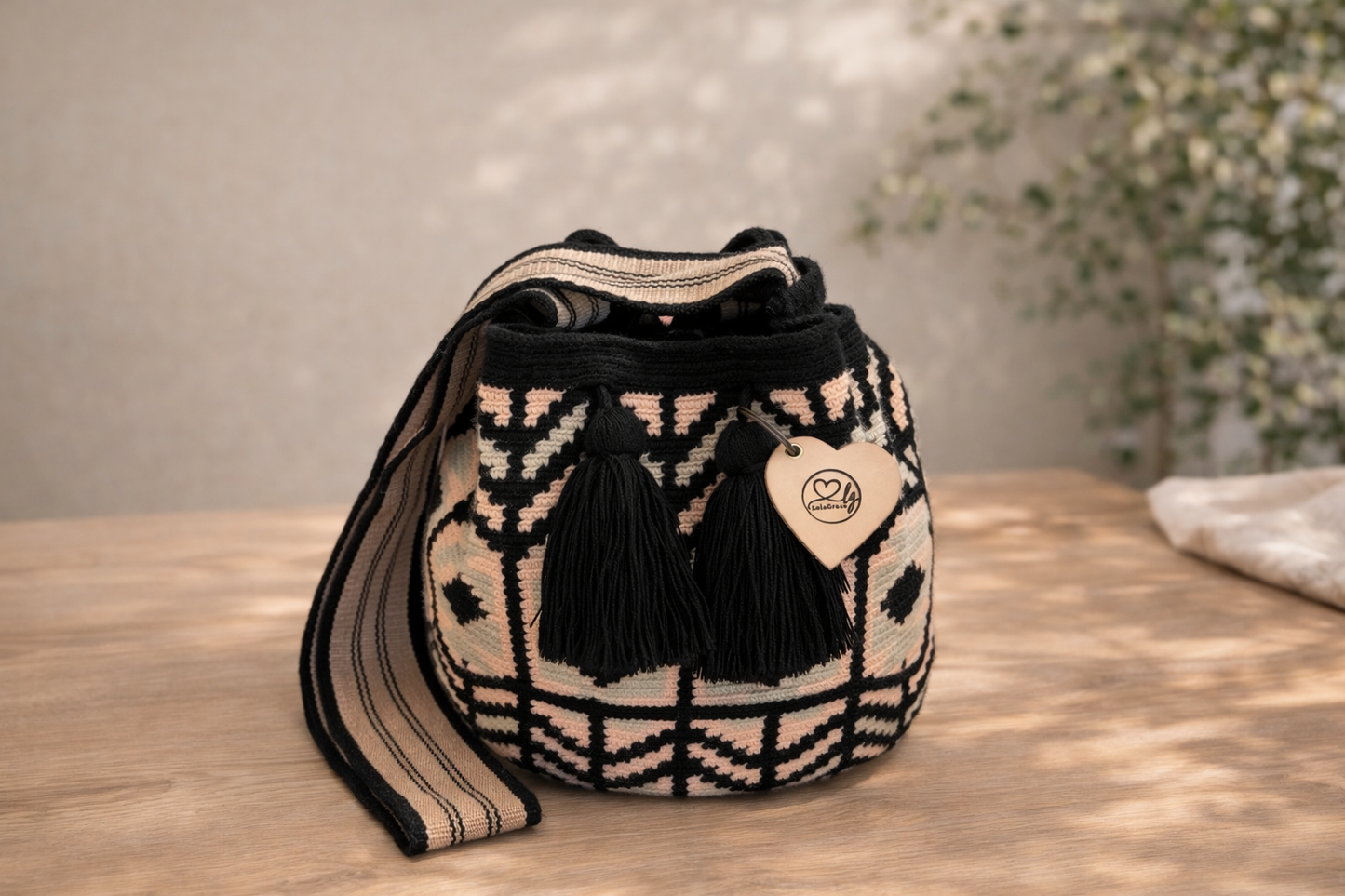 Silvia Large Wayuu mochila tas van LolaGrace op houten tafel, handgemaakt door de Wayuu gemeenschap in La Guajira, Colombia, met zwart en zachtroze patroon en zwarte kwasten.