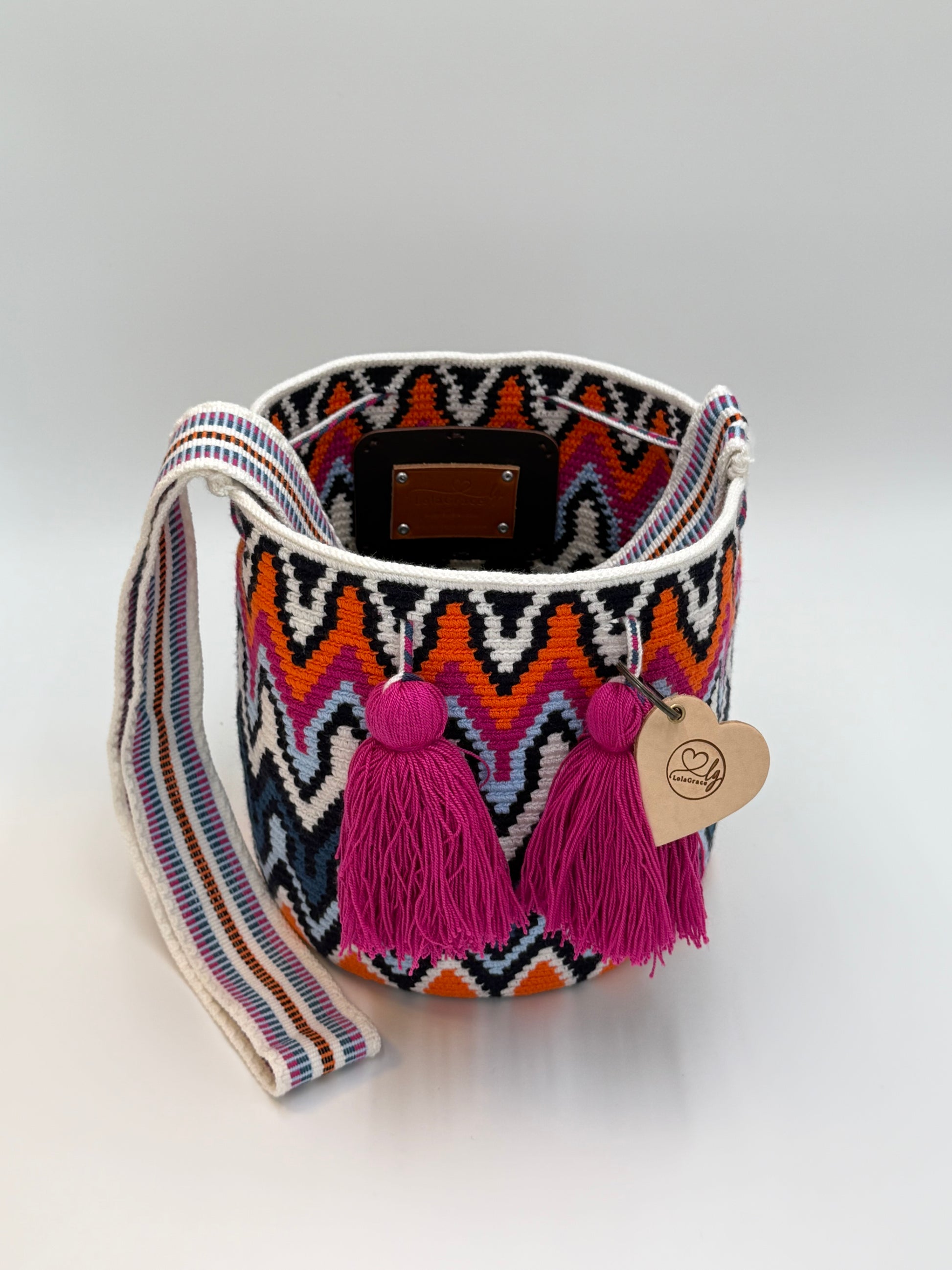 Open Rosaura Medium Wayuu mochila tas van LolaGrace van bovenaf met handgehaakt patroon in zwart, wit, oranje en blauw en roze kwasten.