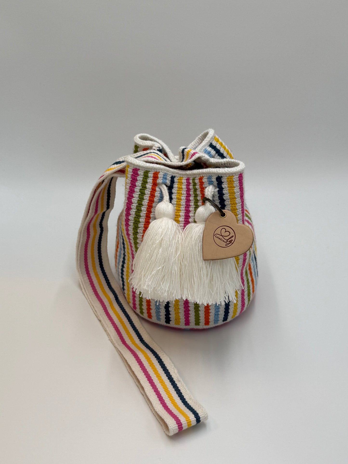 Voorkant van handgemaakte Wayuu tas Catalina Sofia Medium van LolaGrace met kleurrijke verticale strepen in roze, geel, blauw en groen en witte kwastjes.